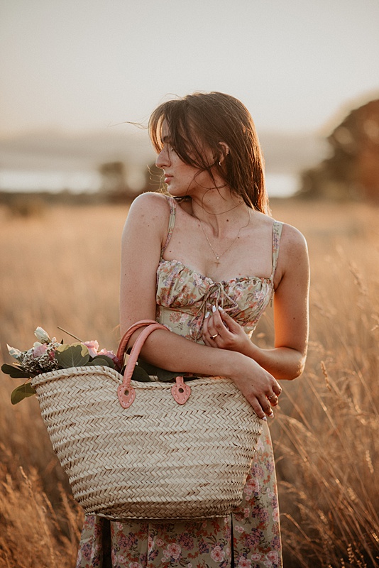 photo of high school senior girl holding farmers market basket with flowers standing in golden field of grass at sunset