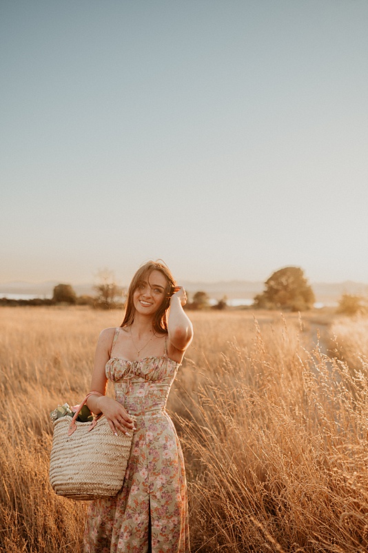 High school senior girl wearing floral dress holding french market basket of flowers walking through tall field of grass at sunset - Seattle senior photo locations top pick
is Discovery Park
