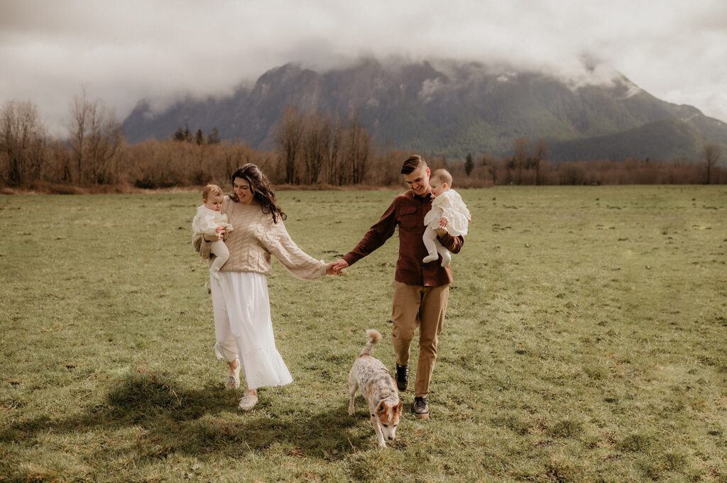 lifestyle photography image of a Couple holding hands while carrying twin baby girls. Dog runs between them as they frolic under Mount Si in North Bend WA