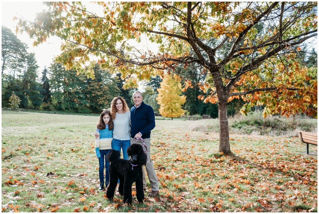 Family of three posing with black poodle under gorgeous tree in fall colors
