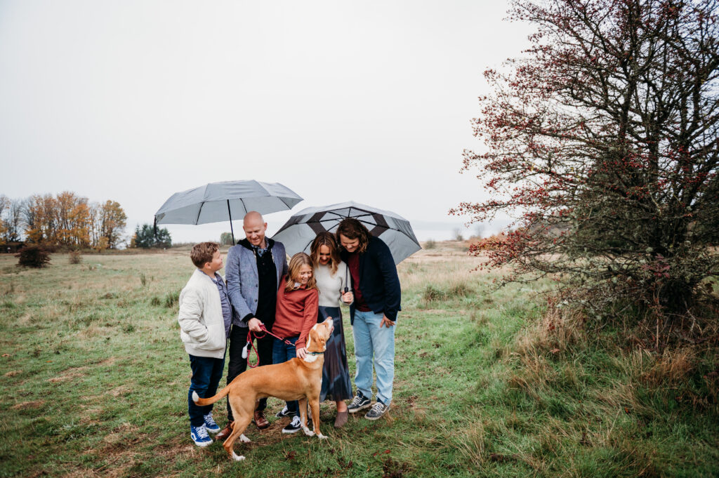 Family of 5 holding umbrellas, smiling and laughing at dog