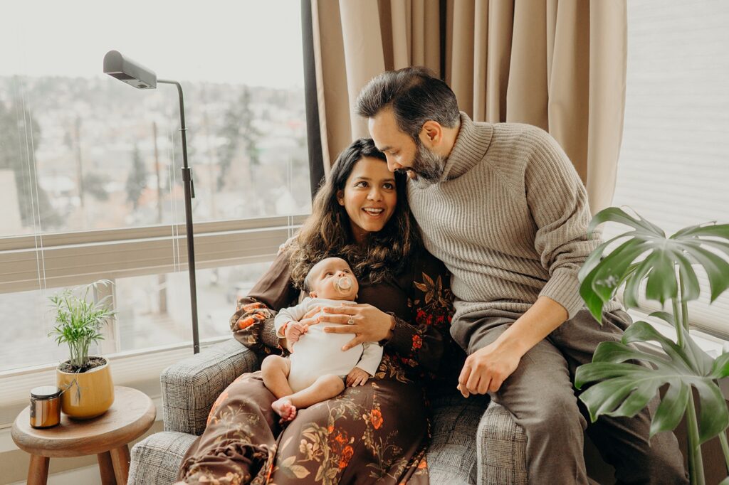 Mom, dad, and newborn baby sitting on chair in front of large window in a aesthetically pleasing home with plants