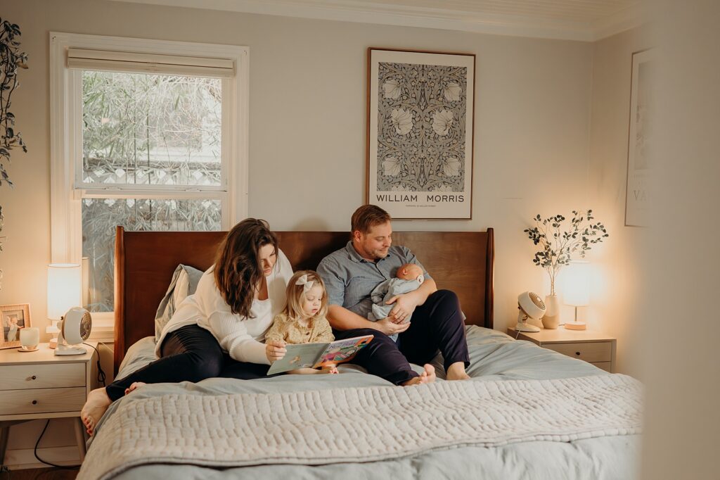 Family sitting on bed reading a book and cuddling newborn baby in cozy bedroom. Their outfits perfectly match the vibe of their home