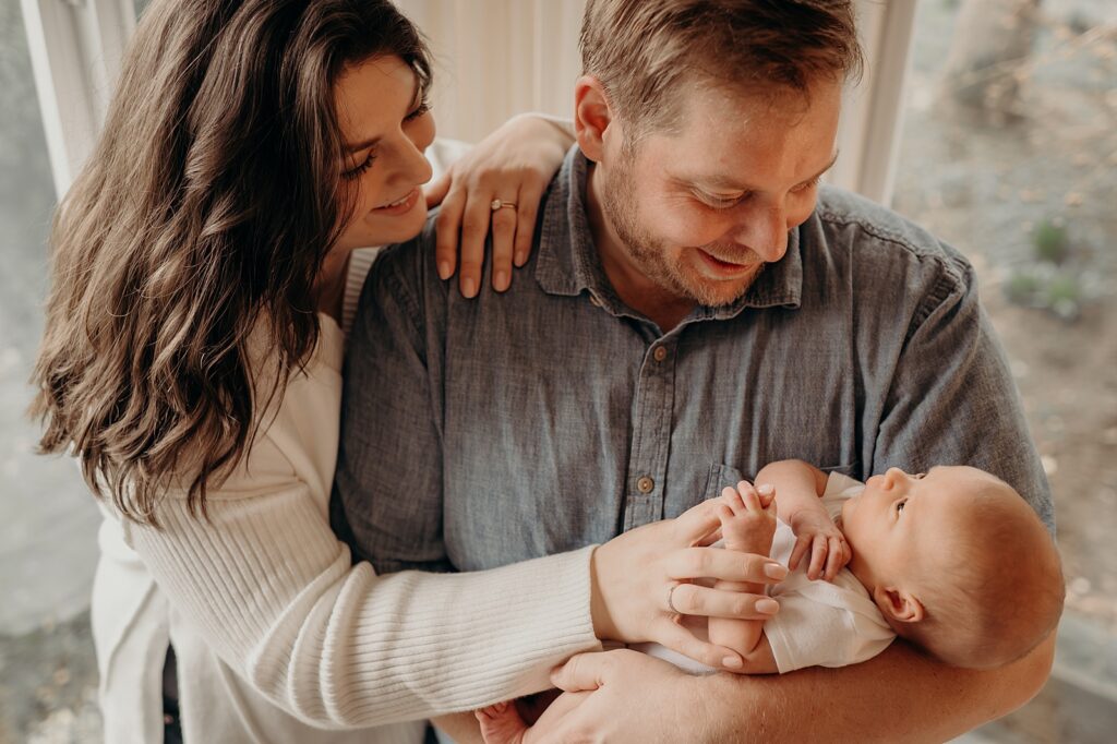 parents smile down at newborn baby standing in front of large living room windows