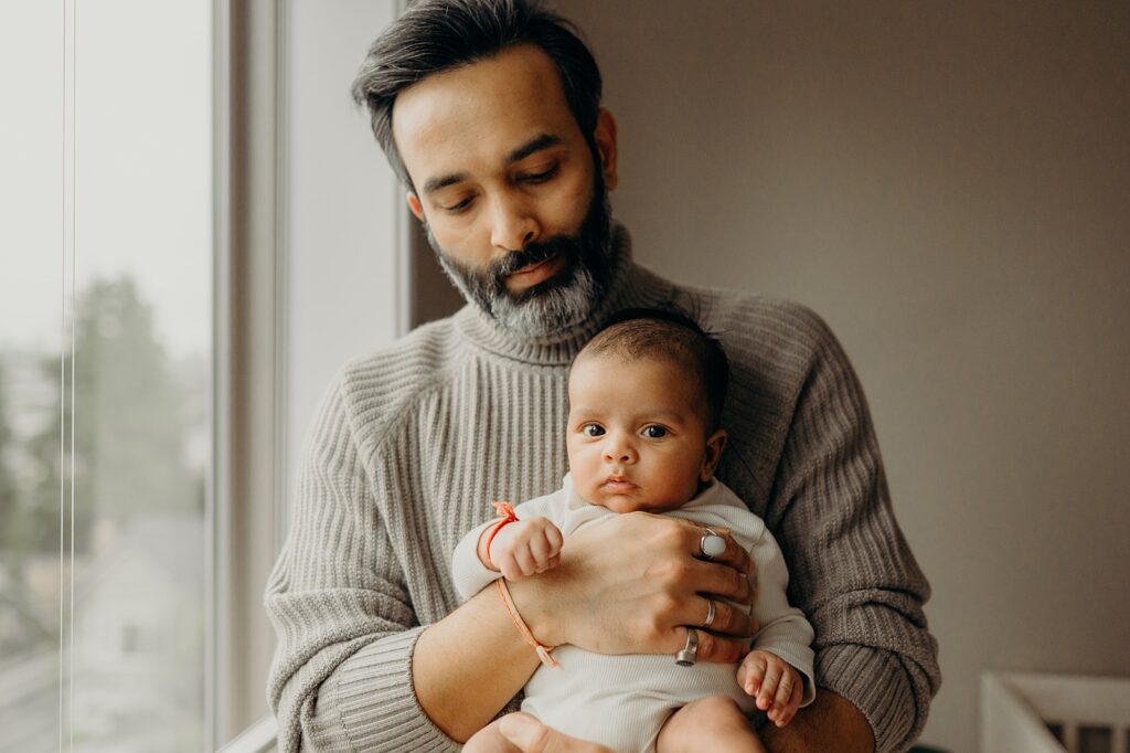 dad holding baby boy looking intently at the camera standing in front of large window